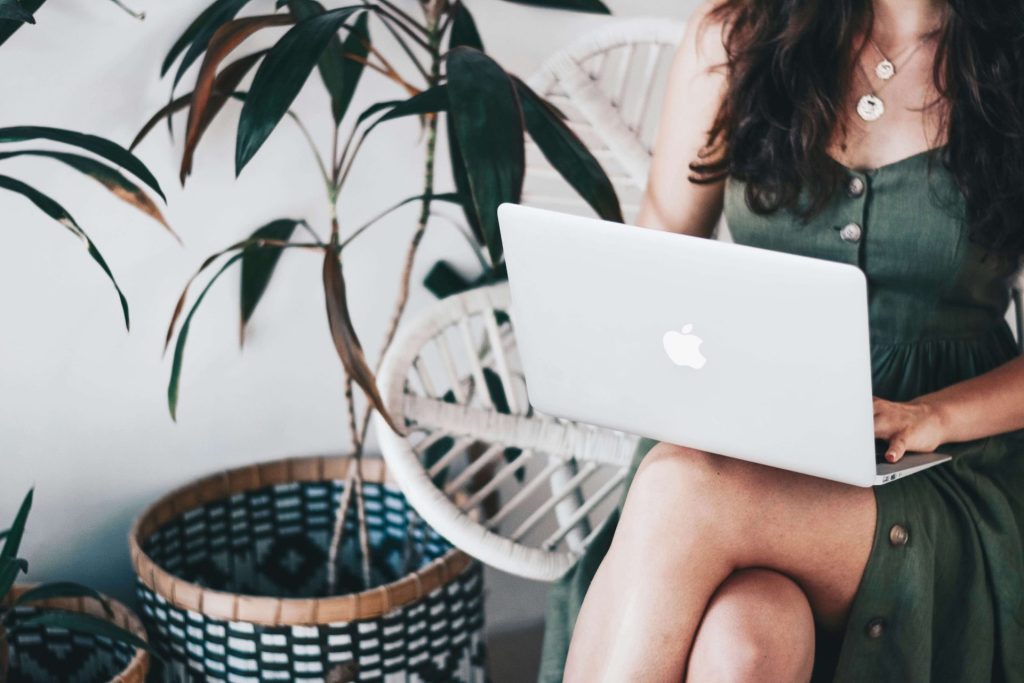Image of a woman wearing a green dress sitting in a chair by a plant holding a laptop. Work with a skilled therapist who can help you with your anxiety, depression, grief, and more with online therapy in Parkland, FL.