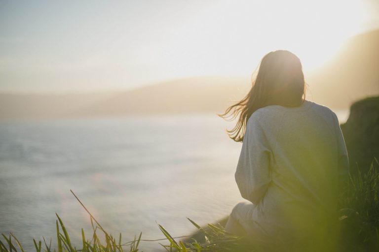 Image of a woman sitting on a cliff near the ocean looking at the water during sunrise. Discover how you can manage your depression due to cheating with the help of depression counseling in Coconut Creek, FL.