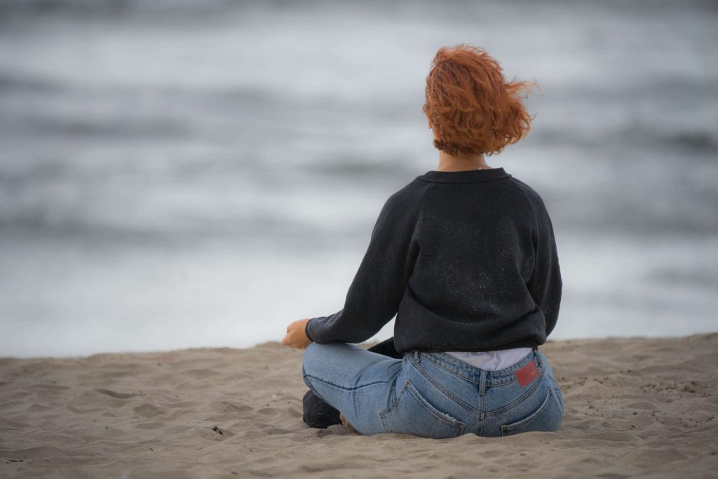 Image of a woman sitting on the beach in the sand looking at the ocean on a cloudy day. Begin finding support for your depression symptoms with the help of depression counseling in Parkland, FL.