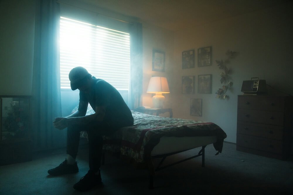 Image of a man sitting on the edge of his bed feeling sad in a dark room. Overcome your suffering and find help with depression counseling in Parkland, FL.