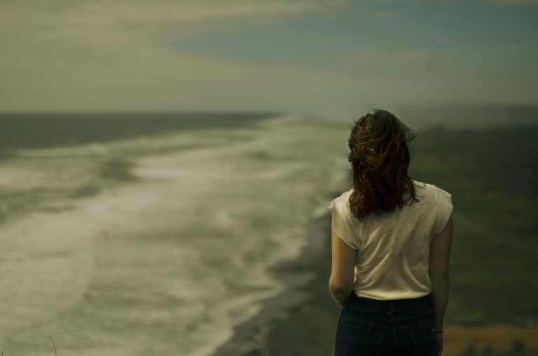 Image of a woman standing on a cliff near a beach on a windy and cloudy day. If you struggle with anxiety due to life transitions, learn how CBT for anxiety in Coconut Creek, FL can help you cope.